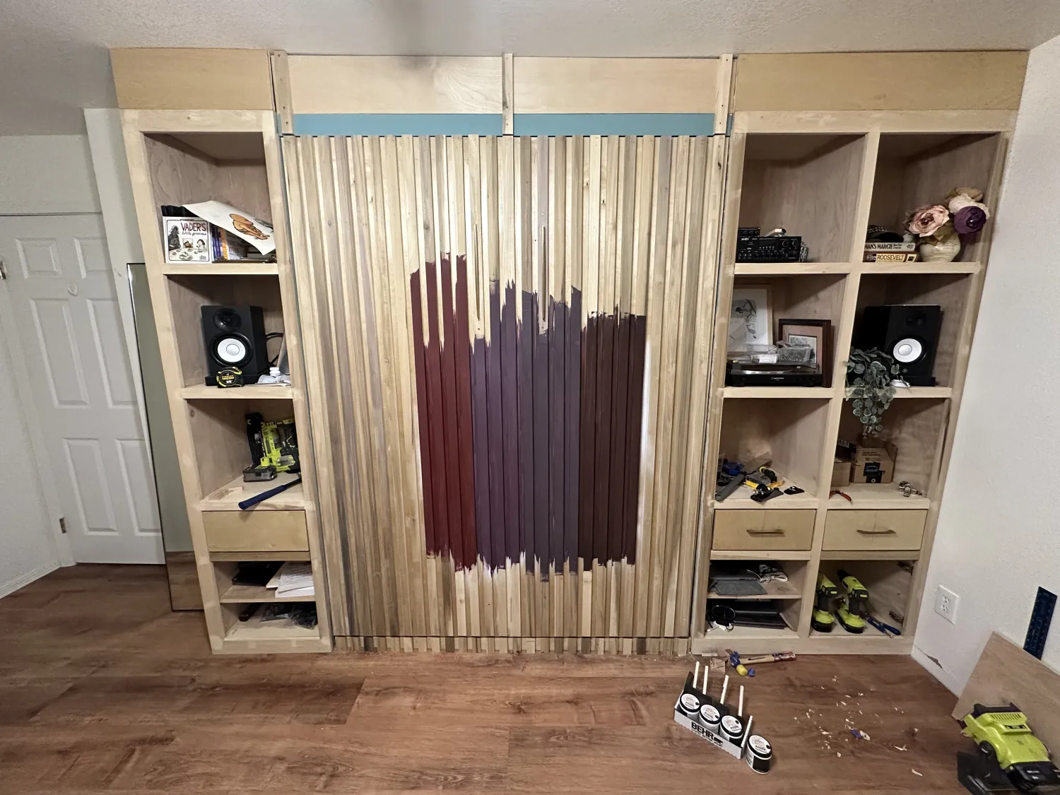 Built-in wall unit and Murphy bed mid-build, raw wood with a stain-test gradient
